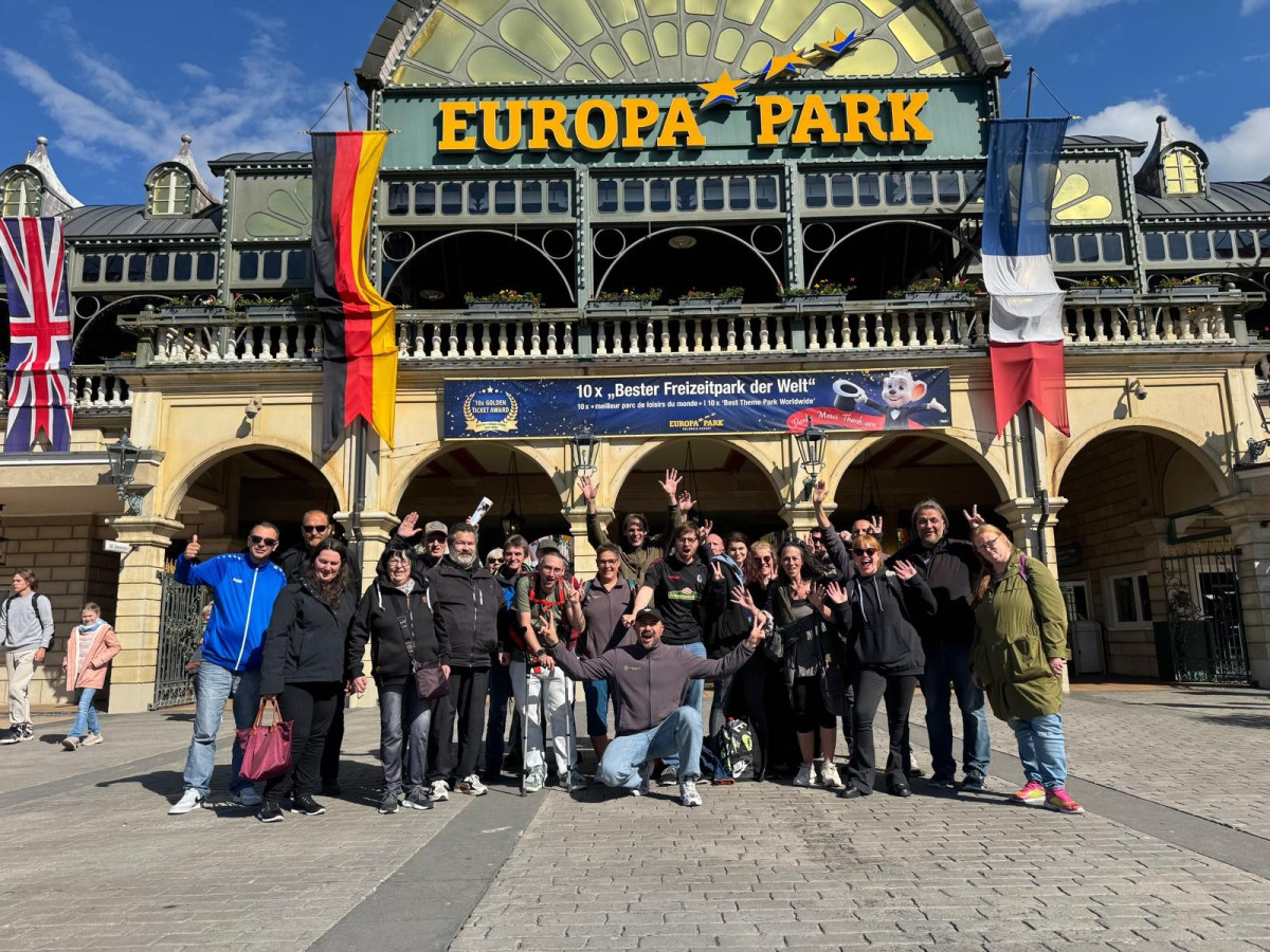Gruppe posiert jubelnd und winkend vor dem Eingang von &bdquo;Europa Park&ldquo; mit L&auml;nderflaggen.