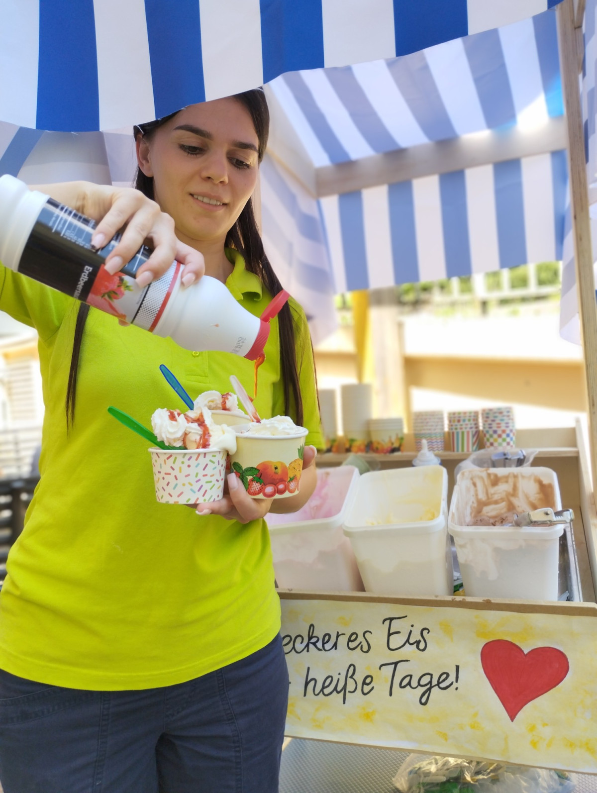 Eine Frau tr&auml;ufelt Schokosauce &uuml;ber zwei Eisbecher an einem Stand mit Schild &bdquo;...eckeres Eis...hei&szlig;e Tage!&ldquo;.