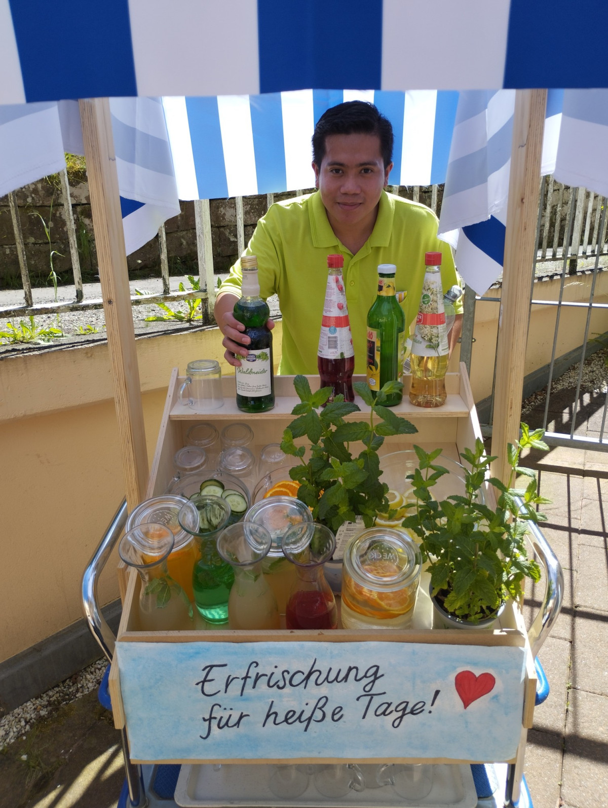 Ein Mann pr&auml;sentiert an einem Getr&auml;nkewagen Flaschen und Sirup, im Schild steht &bdquo;Erfrischung f&uuml;r hei&szlig;e Tage!&ldquo;.