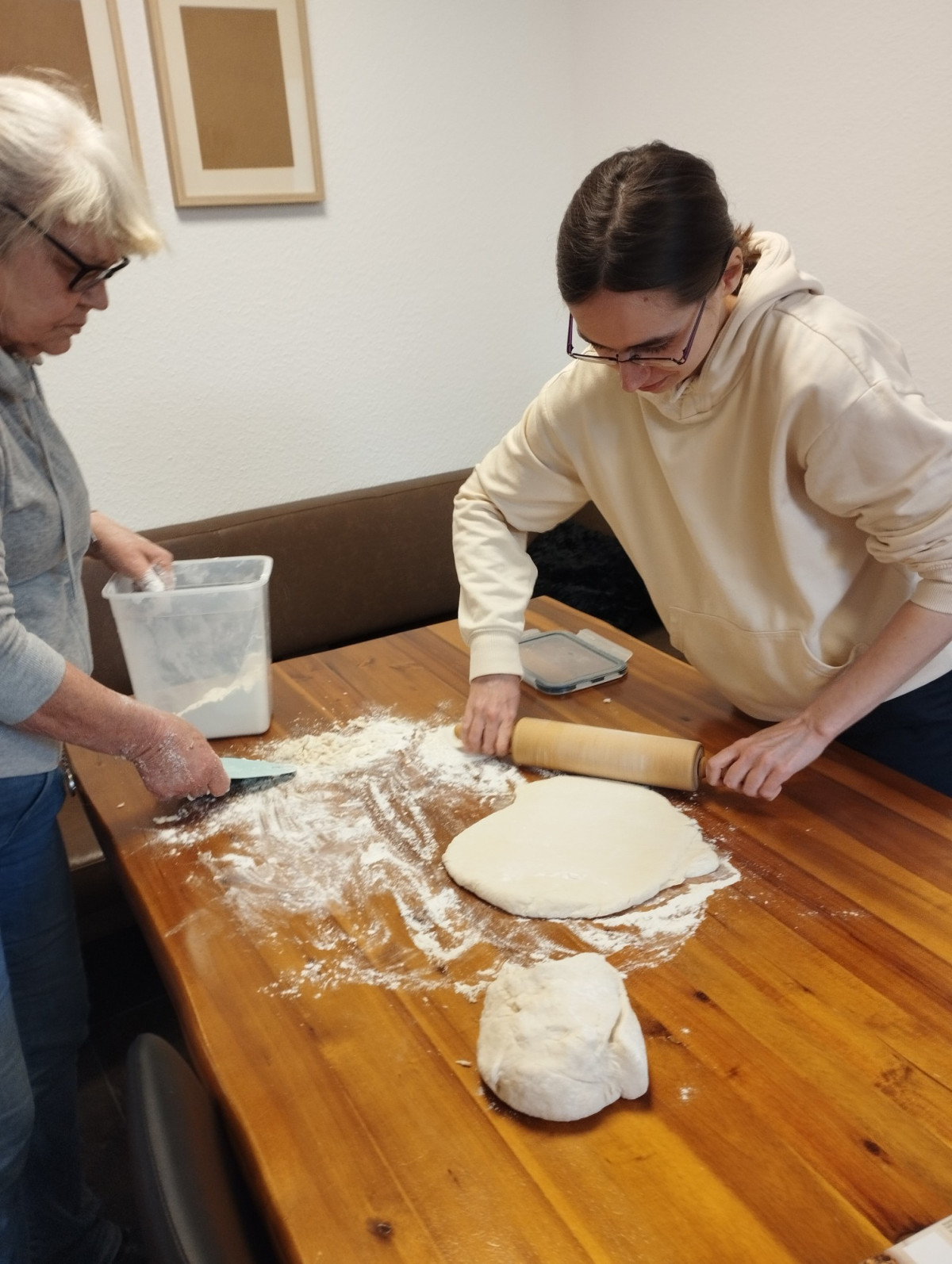 Zwei Personen rollen gemeinsam Teig auf einem mit Mehl best&auml;ubten Holztisch aus.