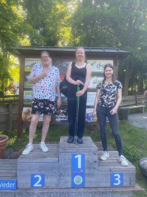 Drei Personen stehen l&auml;chelnd auf einem Siegerpodest mit den Ziffern "1", "2" und "3", eine h&auml;lt einen Minigolfschl&auml;ger.