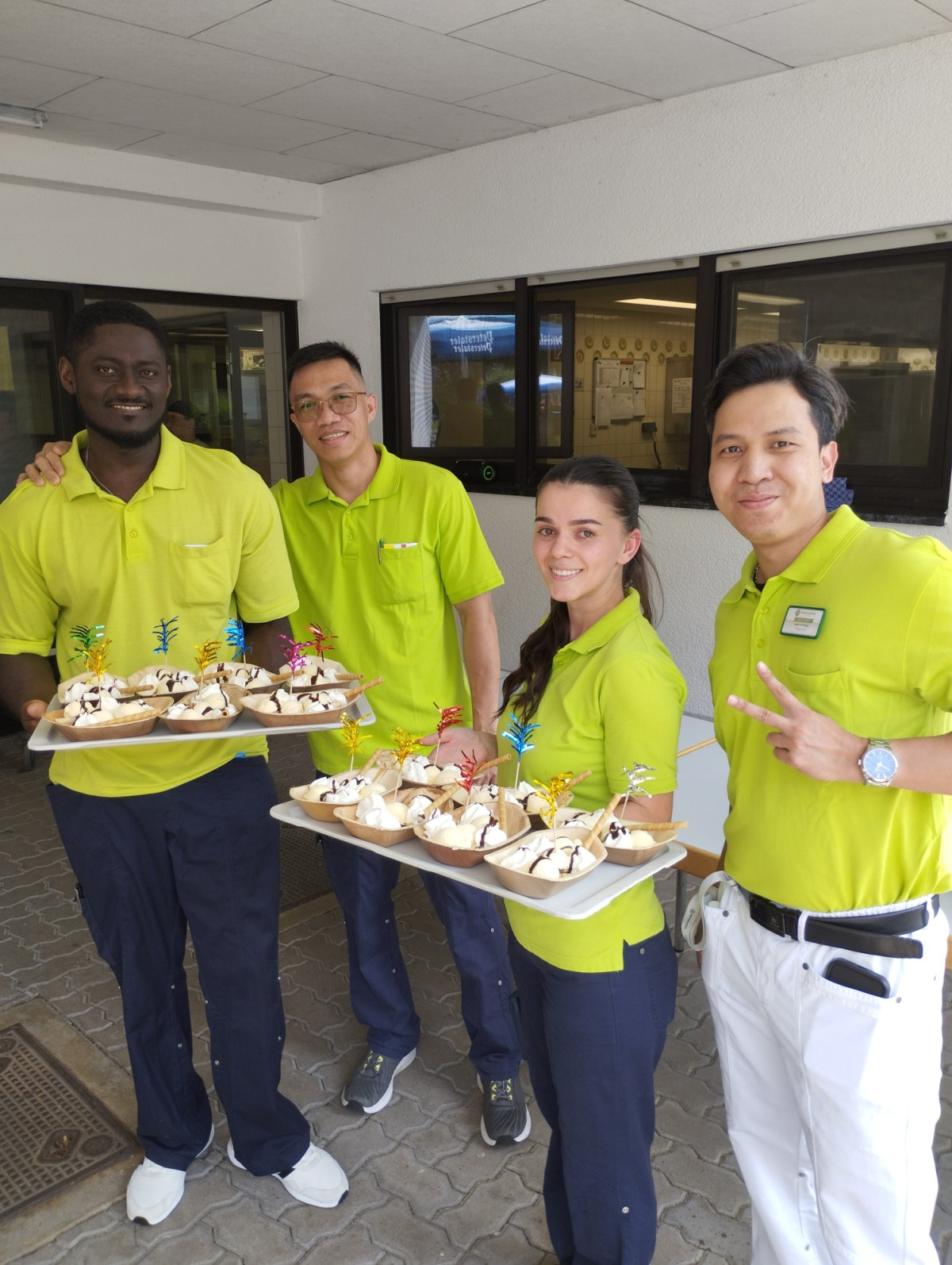 Vier Personen in gelben Poloshirts pr&auml;sentieren l&auml;chelnd Tabletts mit dekorierten Desserts, einer zeigt ein Peace-Zeichen.