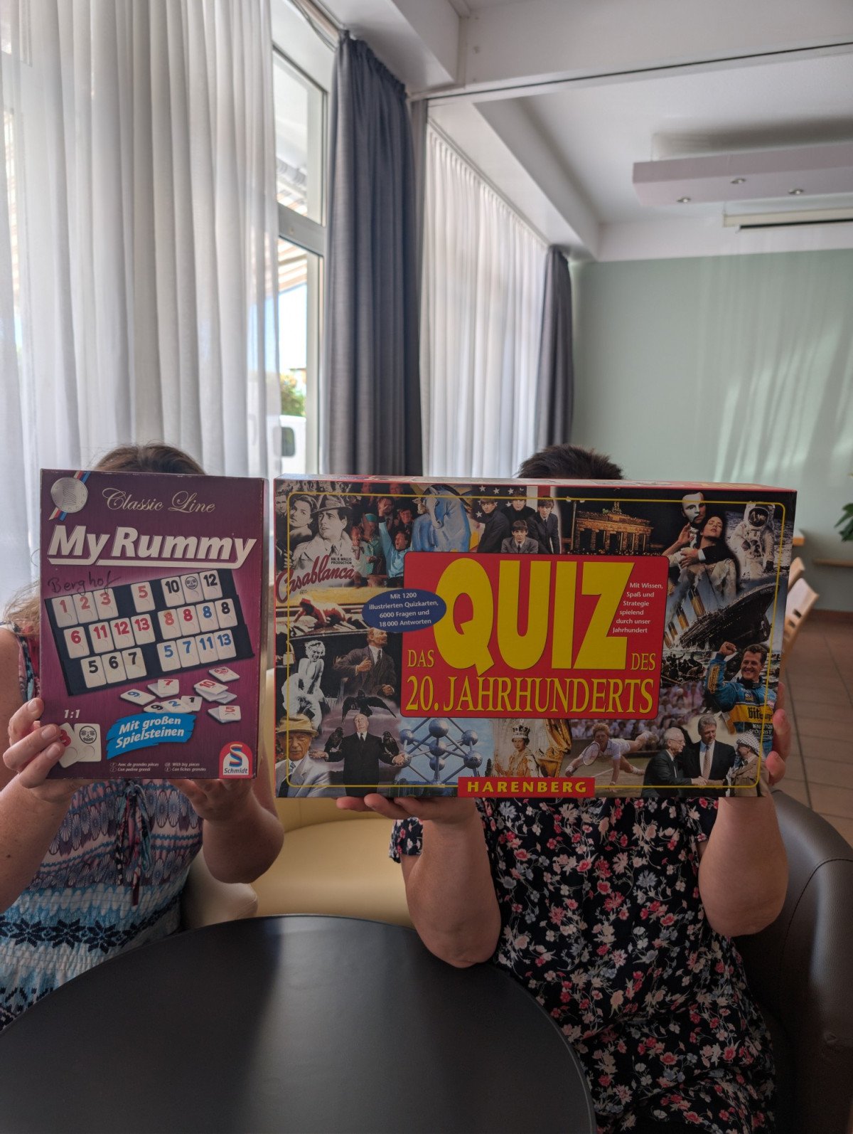 Zwei Personen halten Gesellschaftsspiele hoch, &bdquo;My Rummy&ldquo; und &bdquo;Quiz des 20. Jahrhunderts&ldquo;, die ihre Gesichter verdecken.