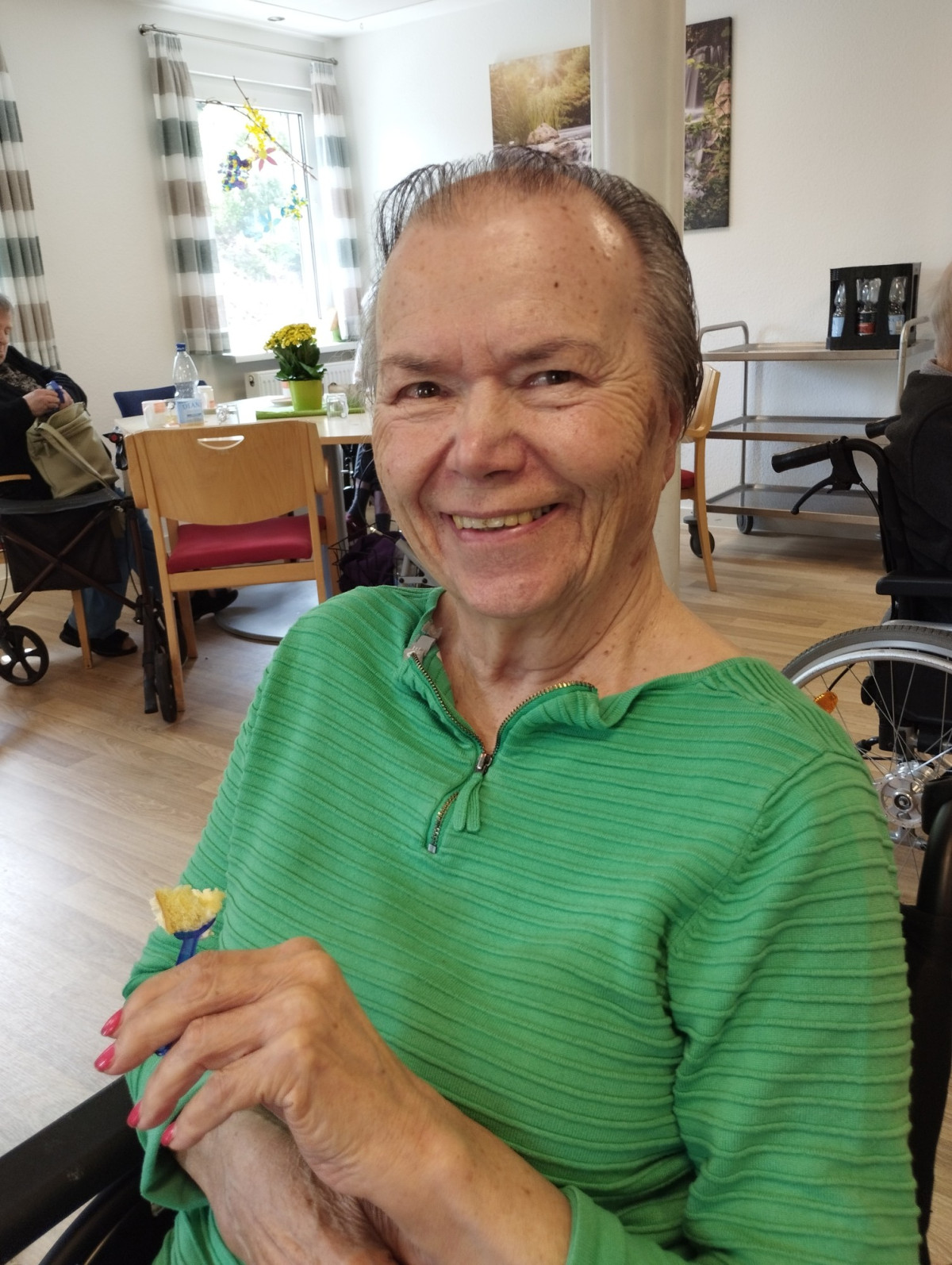 Eine Frau in gr&uuml;nem Pullover l&auml;chelt fr&ouml;hlich in die Kamera und h&auml;lt eine kleine Bl&uuml;te in der Hand.