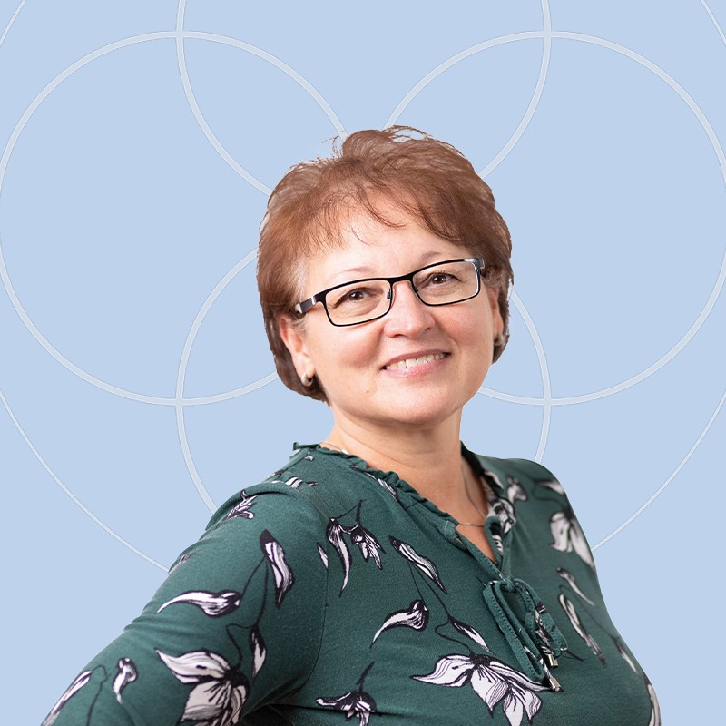 Frau mit r&ouml;tlichen Haaren und Brille in gr&uuml;nem Oberteil vor blauem Hintergrund.