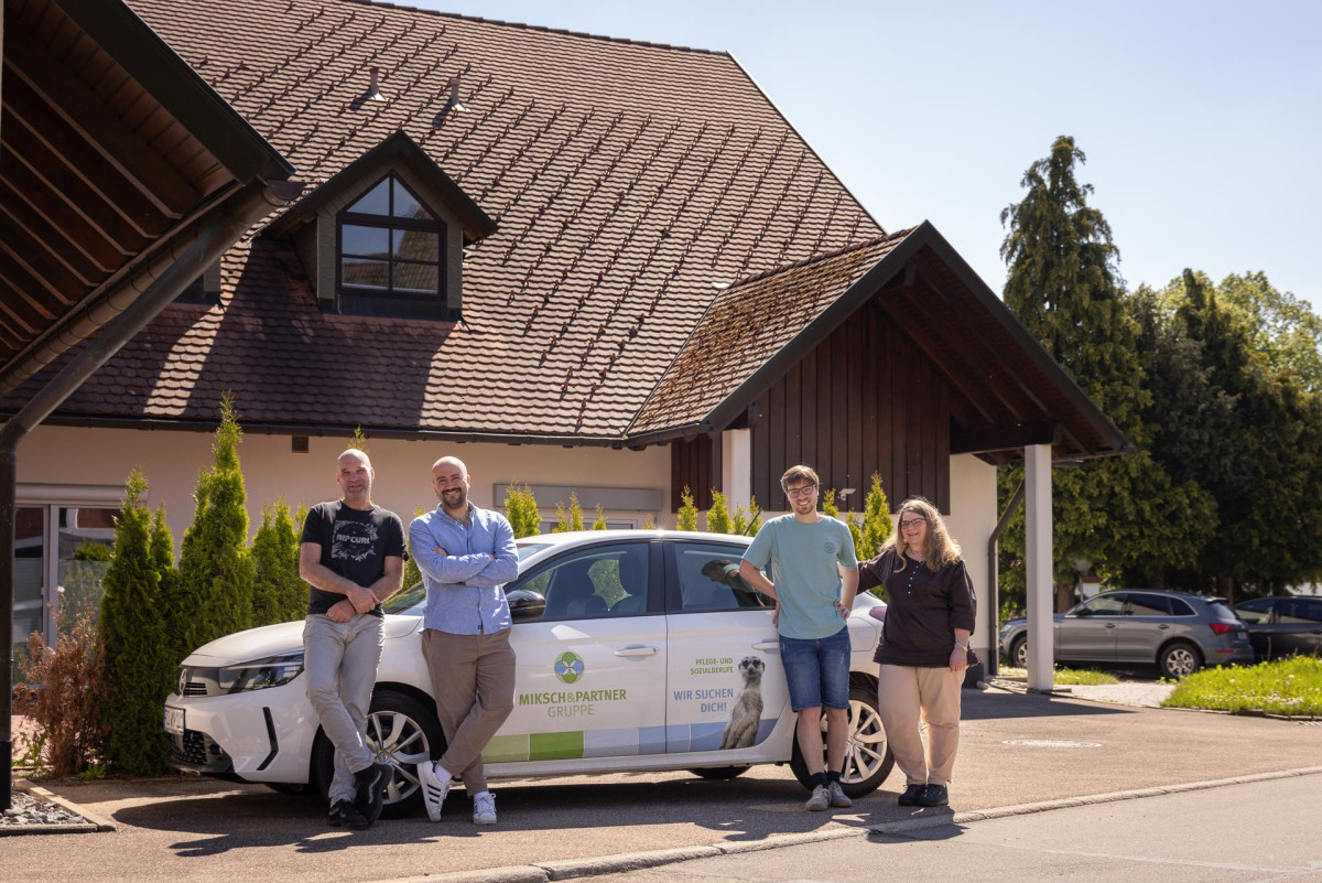F&uuml;nf Personen stehen neben einem wei&szlig;en Auto vor einem Haus.
