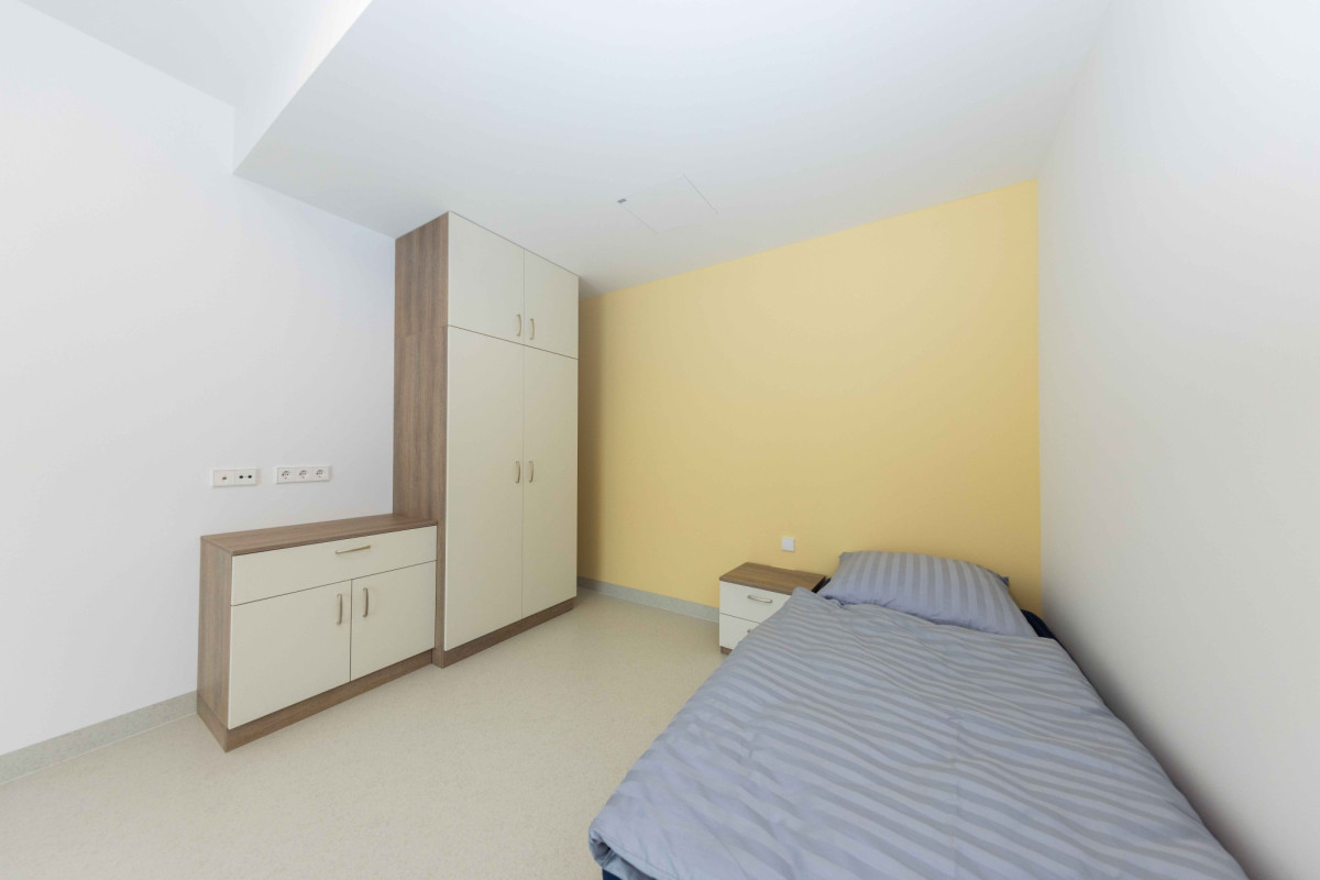 Kleines Zimmer mit Bett, Schrank und gelblicher Wand.
