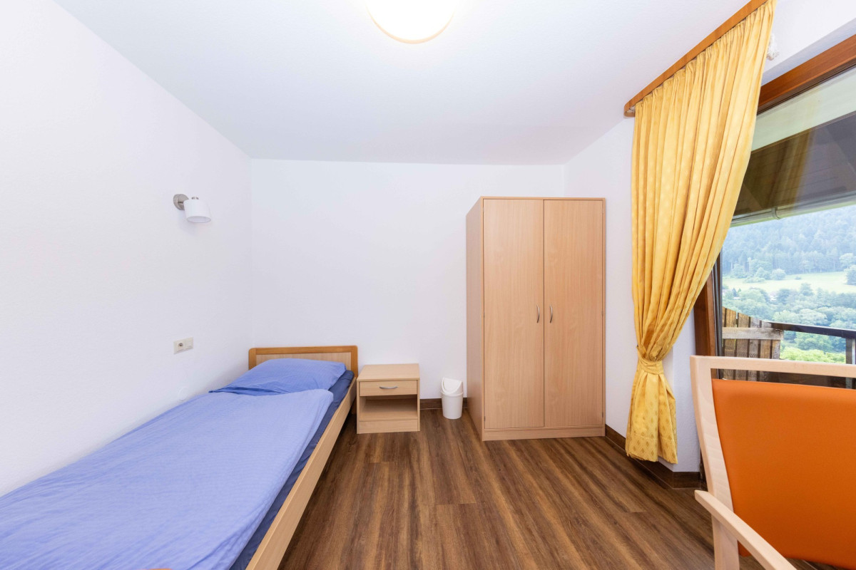 Einfaches Schlafzimmer mit Einzelbett, Kleiderschrank und Fenster.