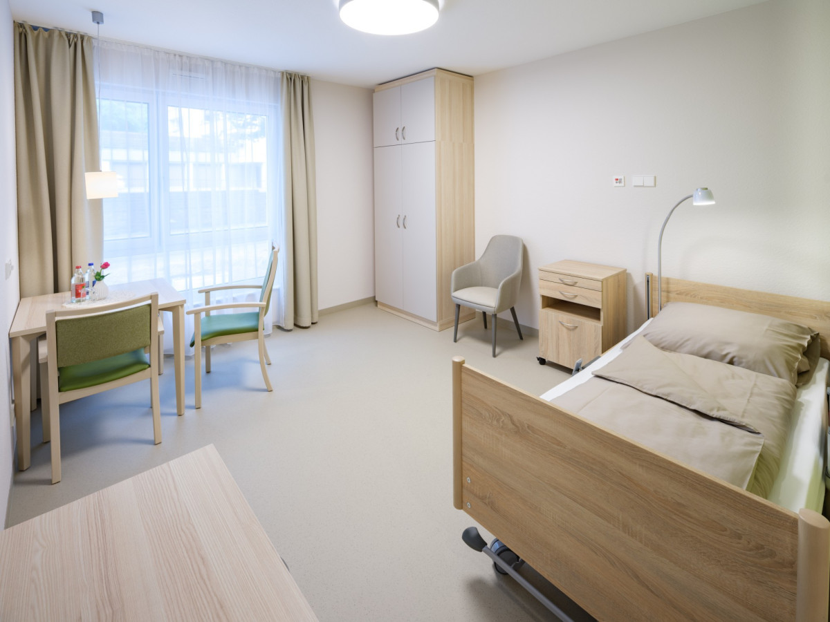 Helles Pflegezimmer mit Bett, Schreibtisch und Sitzbereich.