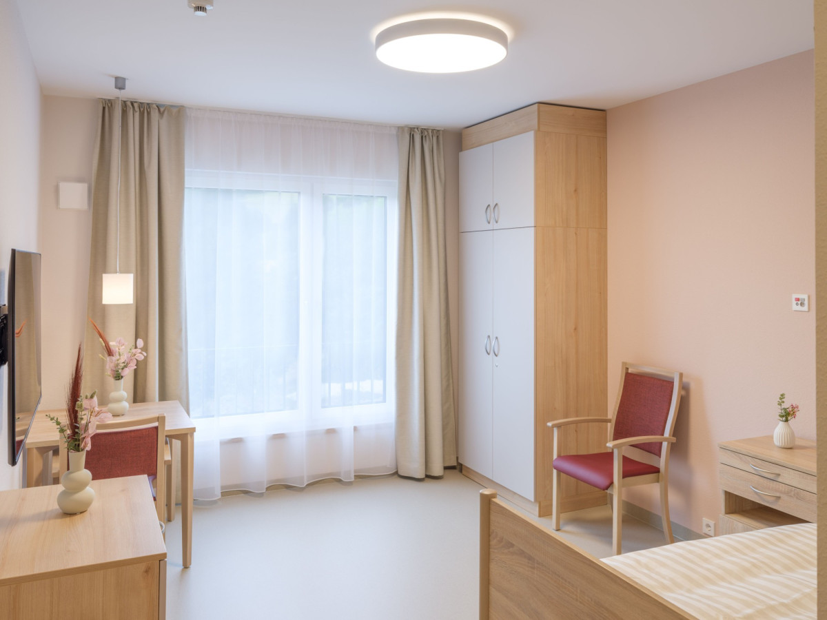 Helles Seniorenzimmer mit Bett, Schrank und Fenster.