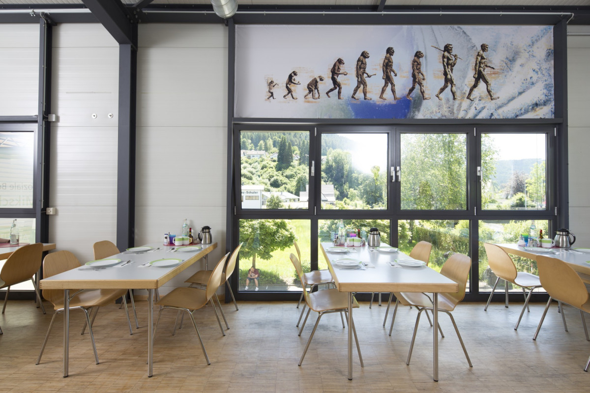 Heller Speiseraum mit Holztischen und St&uuml;hlen, gro&szlig;en Fenstern mit Bergblick und Evolution-Kunstwerk an der Wand.